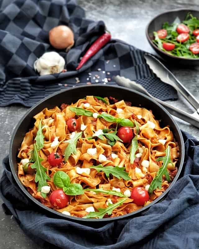 One Pot Pasta mit Peperoni, Rucola und Mozzarella in der Pfanne fotografiert. Im Hintergrund steht eine schale mit frischem Salat. Daneben auf einem Küchenhandtuch liegt eine Zwiebel, eine Knoblauchzehe und eine Peperoni.