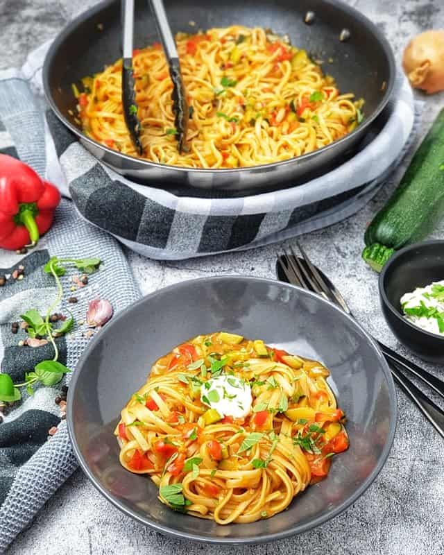 One Pot Pasta mit Paprika und Zucchini in einer grauen Schale angerichtet. Im Hintergrund eine Pfanne in der das Gericht gekocht wurde.